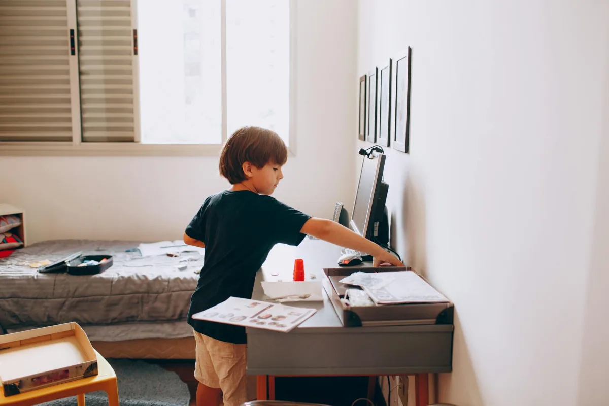 A child at a small desk in a bedroom doing homework with the door closed, late afternoon light through one window, a CO₂ reading visible on a phone screen on the desk.