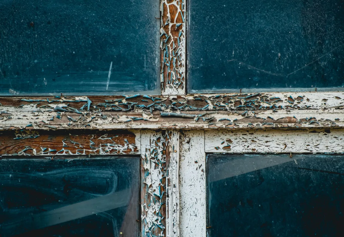 A peeling painted window sill in an older home with an EPA Lead-Safe Certified Renovation logo visible on a contractor sign in the foreground.