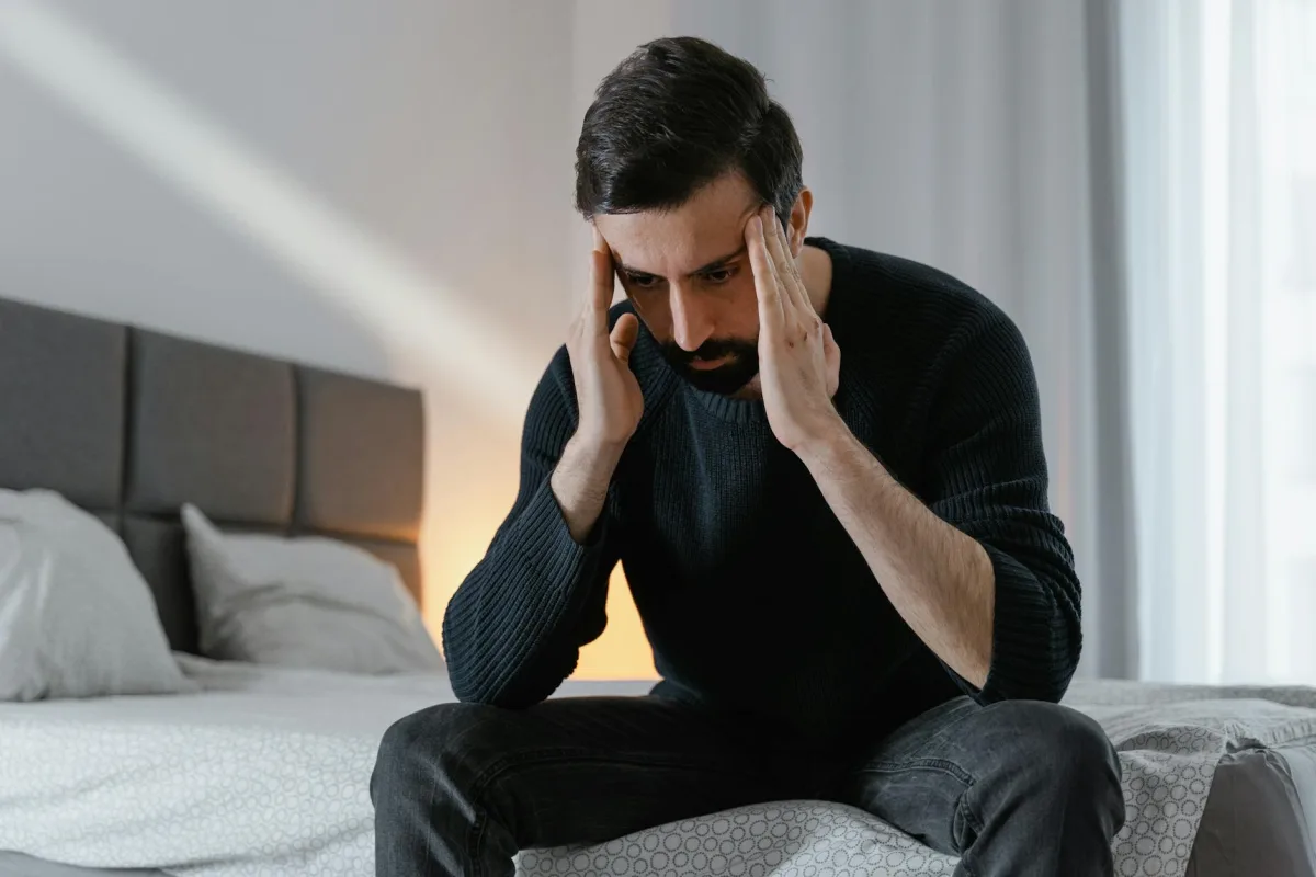 A person resting in a darkened bedroom with a phone showing a migraine-tracking app next to a Terrestream sensor reading.