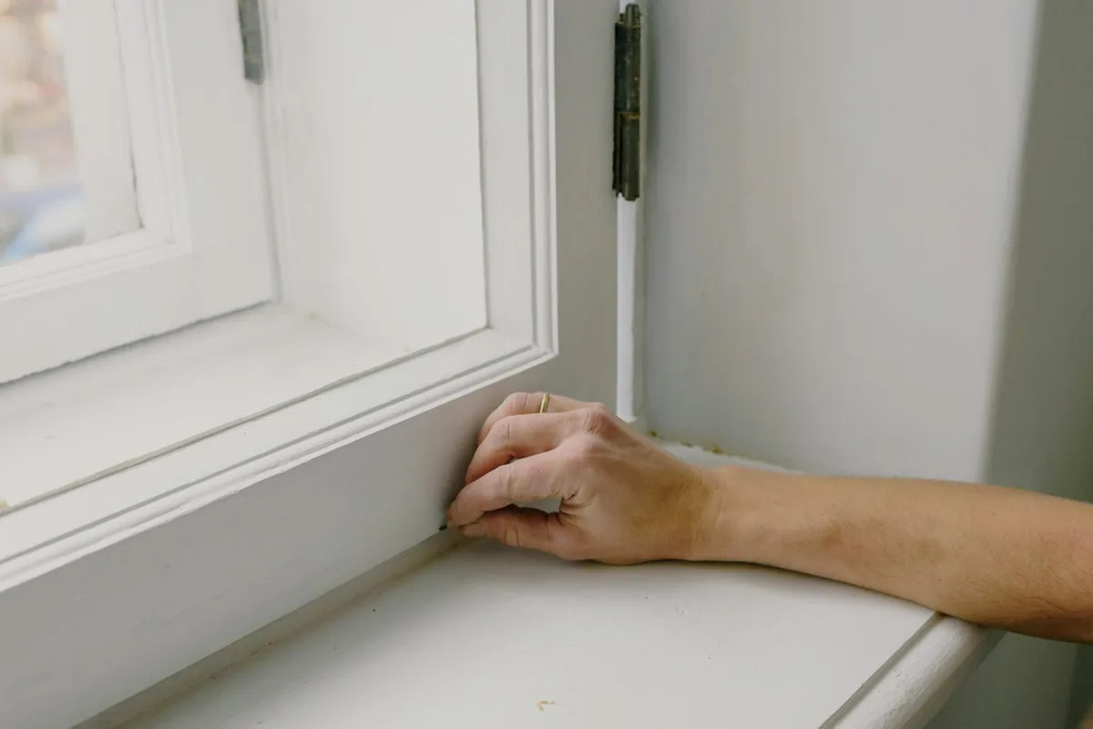 Une main gantée essuyant une mince couche de cendre pâle d'un rebord de fenêtre avec un chiffon microfibre humide, avec un aspirateur HEPA au sol derrière.
