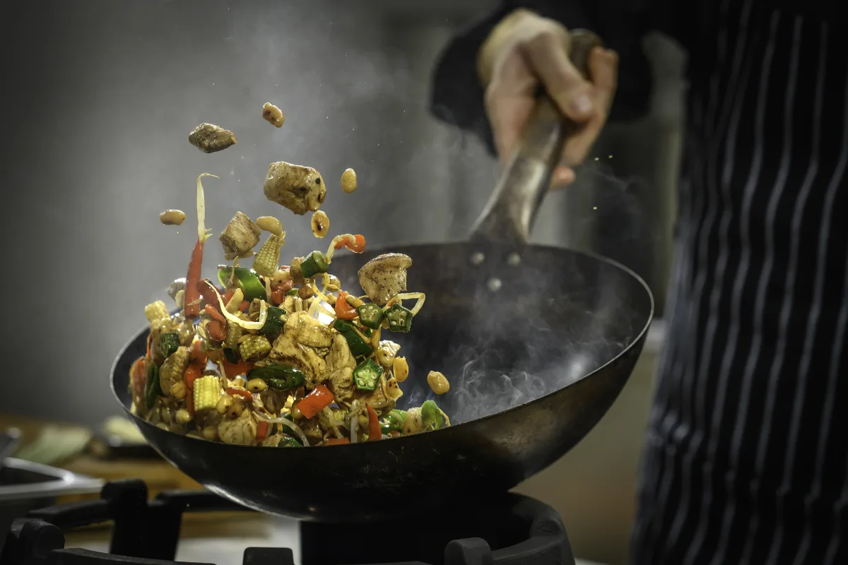 A chef tossing peanuts in a wok over a gas burner, steam and aromatics rising
