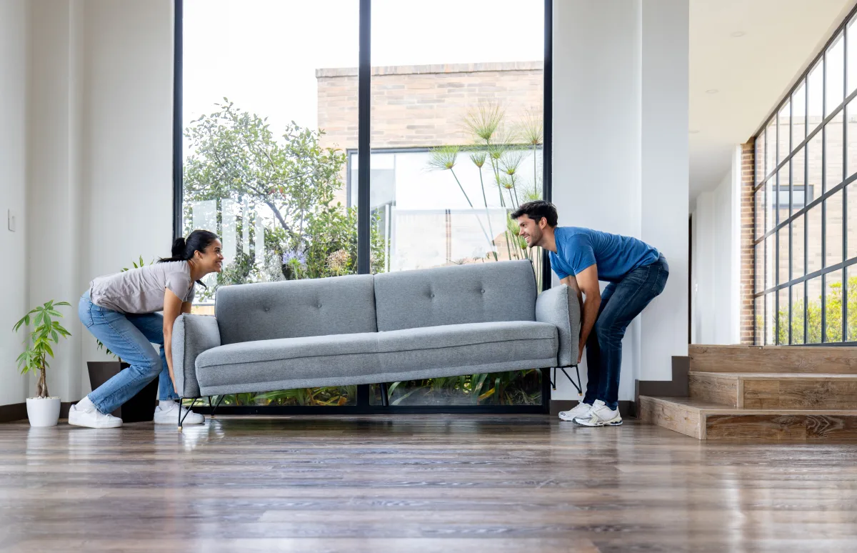 A couple carrying a new gray sofa into a sunlit living room